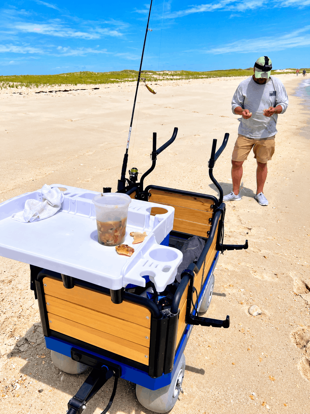 A motorized beach fishing cart with a fillet tray, wood-grain sides, and sand spike holders shown on a sandy beach with a person standing in the background near a fishing rod.